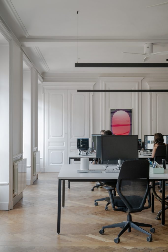 Open space avec de grand bureaux et chaises ergonomiques. Longs luminaires noirs en led au plafond.